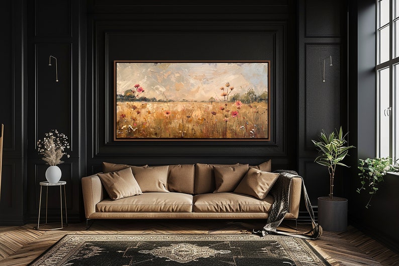 May include: A painting of a field of wildflowers in shades of pink, yellow, and green, with a cloudy sky in the background. The painting is framed and hanging on a black wall in a room with a brown leather sofa and a potted plant.