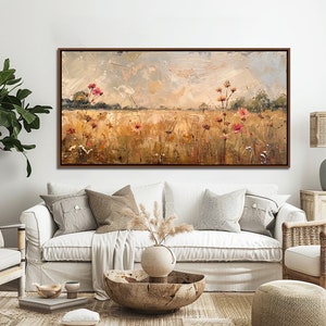 May include: A painting of a field of wildflowers in shades of pink, yellow, and green, with a cloudy sky in the background. The painting is framed in a brown wood frame and is hanging on a white wall in a living room setting.
