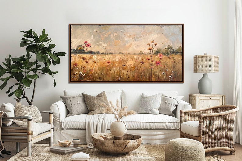 May include: A painting of a field of wildflowers in shades of pink, yellow, and green, with a cloudy sky in the background. The painting is framed in a brown wood frame and is hanging on a white wall in a living room setting.