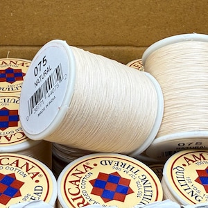 May include: Close-up of several spools of natural-colored quilting thread. One spool is in focus, displaying the number "075" and the word "NATURAL" on its label. The thread is labeled "Americana Glaced Cotton".