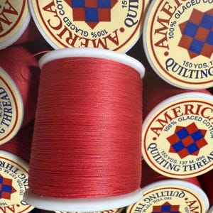 May include: Close-up of red quilting thread spools. The thread is wound on white spools, with labels that read "Americana 100% Glaced Cotton Quilting Thread." Each spool contains 150 yards of thread.