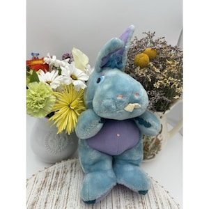 Könnte beinhalten: Ein blaues Plüschkaninchen mit lila Bauch und Akzenten. Das Kaninchen hat lange Ohren, eine kleine rosa Nase und einen einzelnen Zahn. Es steht vor einer weißen Vase mit bunten Blumen und einer Vase mit getrockneten Blumen.