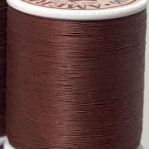 Puede incluir: Un carrete de hilo marrón oscuro, enrollado alrededor de un núcleo de cartón blanco. La parte superior del carrete presenta una etiqueta circular con texto y un diseño geométrico en rojo, azul y blanco. El hilo parece liso, para coser o manualidades.