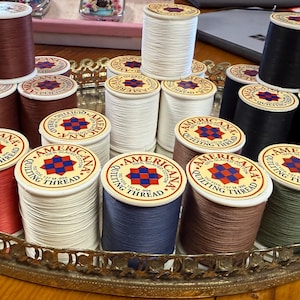 May include: A collection of spools of Americana quilting thread in various colors, including red, white, blue, brown, and black. The spools are arranged on a decorative silver tray. Each spool has a label with a red, white, and blue star design.