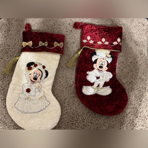 Op de afbeelding: Twee kerstsokken met Disney-personages. Eén is wit met Minnie Mouse in een trouwjurk, de andere is rood met Mickey Mouse in een smoking. Beide hebben gouden kwastjes en rode fluwelen tops met gouden strikken.