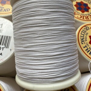May include: Close-up of a spool of white thread, tightly wound around a cream-colored plastic core. The thread is smooth and appears to be cotton. Other spools of thread are visible in the background, with labels that include text such as "American Thread" and "Quilting Thread."