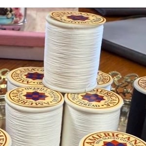 May include: A collection of white thread spools with circular labels. The labels read "Americana Quilting Thread" and feature a starburst design. The spools are stacked on a decorative tray, showcasing the quilting thread.