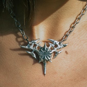 Op de afbeelding: Een zilveren ketting met een grote, ingewikkelde hanger met een stekelig, geometrisch ontwerp. De hanger is bevestigd aan een zilveren ketting.