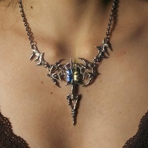 May include: A silver-toned statement necklace with a detailed, ornate design. The necklace features a central pendant with a blue and yellow stone, and is worn against a dark brown top.
