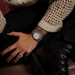 Puede incluir: Un reloj plateado con una esfera blanca y una pulsera de cadena plateada. El reloj est&aacute; en la mu&ntilde;eca de una persona y la pulsera est&aacute; en la misma mu&ntilde;eca. La persona lleva pantalones negros y un su&eacute;ter de crochet blanco.