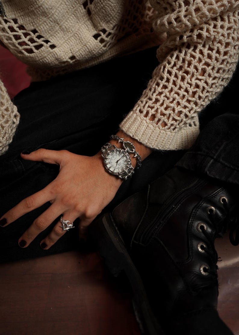 Puede incluir: Un reloj plateado con una esfera blanca y una pulsera de cadena plateada. El reloj est&aacute; en la mu&ntilde;eca de una persona y la pulsera est&aacute; en la misma mu&ntilde;eca. La persona lleva pantalones negros y un su&eacute;ter de crochet blanco.