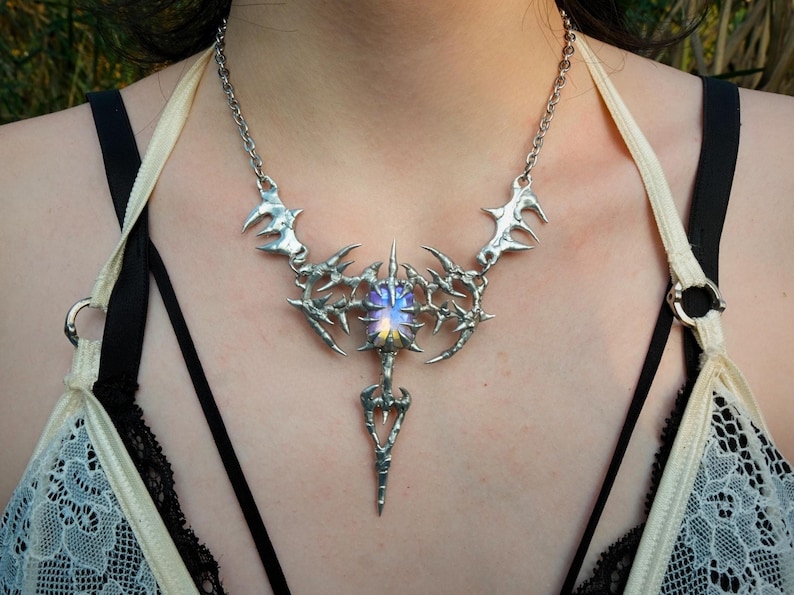 May include: A silver-toned statement necklace with a spiky, ornate design. The pendant features a central, iridescent stone and a long, pointed drop. The necklace is worn against a background of skin and a black and white lace top.