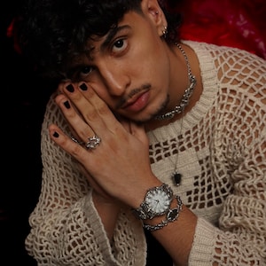 Puede incluir: Una persona lleva un su&eacute;ter de crochet blanco con un reloj plateado, un collar de cadena plateada con un colgante negro y un anillo plateado en el dedo. Tiene esmalte de u&ntilde;as negro en las u&ntilde;as.