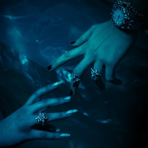 Puede incluir: Dos manos con esmalte de u&ntilde;as oscuro y anillos de plata, una mano lleva un reloj de plata con un dise&ntilde;o detallado, las manos se acercan la una a la otra sobre un fondo azul oscuro.