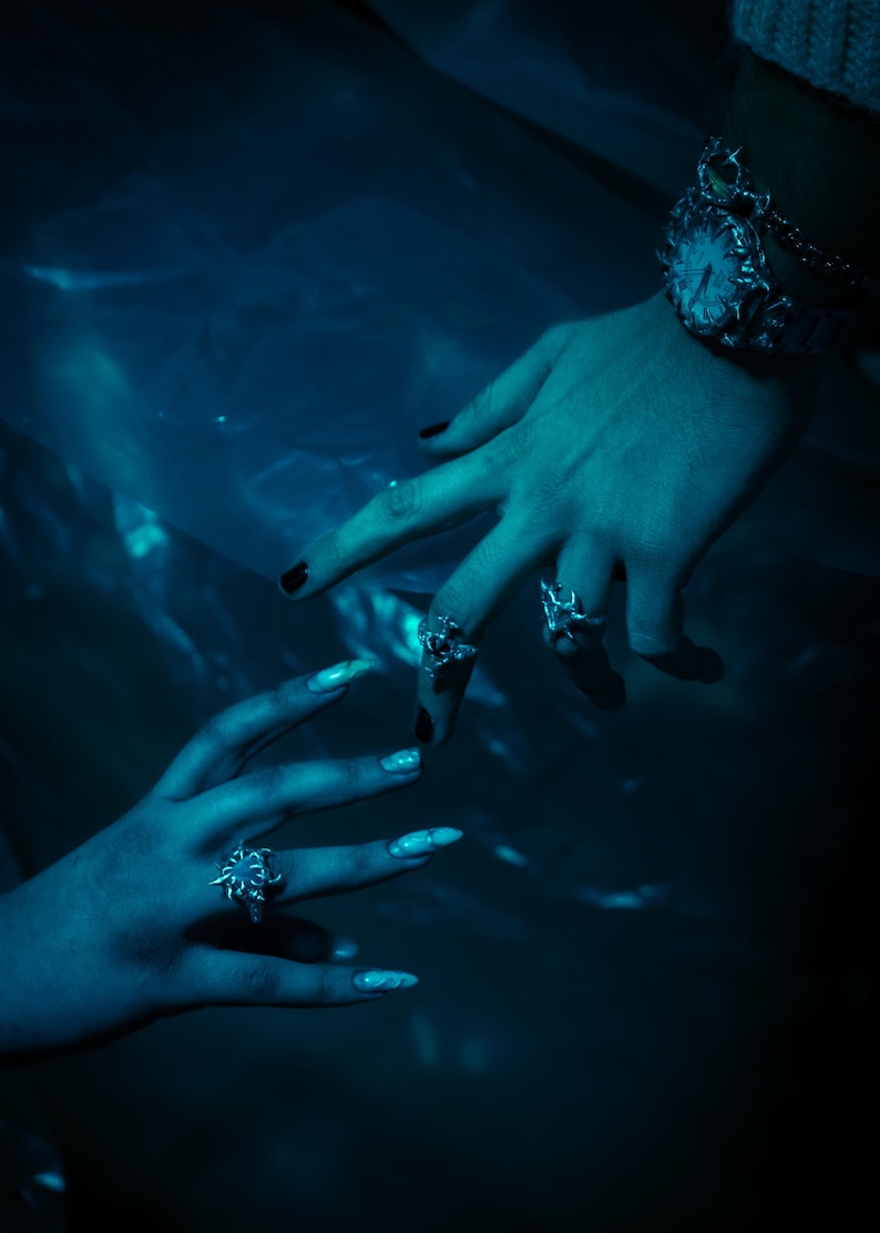 Puede incluir: Dos manos con esmalte de u&ntilde;as oscuro y anillos de plata, una mano lleva un reloj de plata con un dise&ntilde;o detallado, las manos se acercan la una a la otra sobre un fondo azul oscuro.