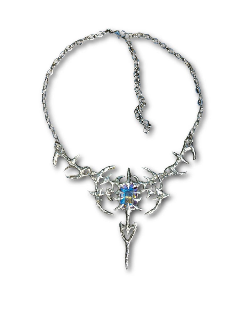 May include: A silver-toned necklace with an intricate, spiky design. The pendant features a central, multi-colored gemstone and a long, pointed drop. The necklace has a chain closure.