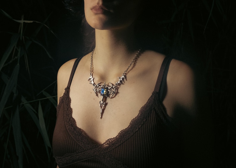 May include: Silver-toned statement necklace with a central blue gemstone and intricate, stylized metalwork. The pendant has a long, pointed drop. The necklace is worn over a brown camisole.