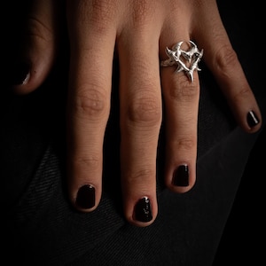 Puede incluir: Un anillo de plata con un dise&ntilde;o en forma de coraz&oacute;n con bordes afilados y puntiagudos. El anillo se lleva en un dedo con esmalte de u&ntilde;as negro.