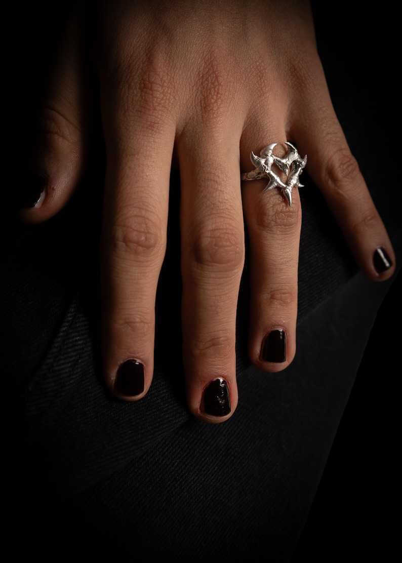 Puede incluir: Un anillo de plata con un dise&ntilde;o en forma de coraz&oacute;n con bordes afilados y puntiagudos. El anillo se lleva en un dedo con esmalte de u&ntilde;as negro.