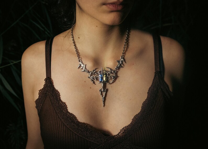 May include: A silver-toned statement necklace with a detailed, ornate design. The necklace features a central pendant with a blue and yellow stone, and is worn against a dark brown top.