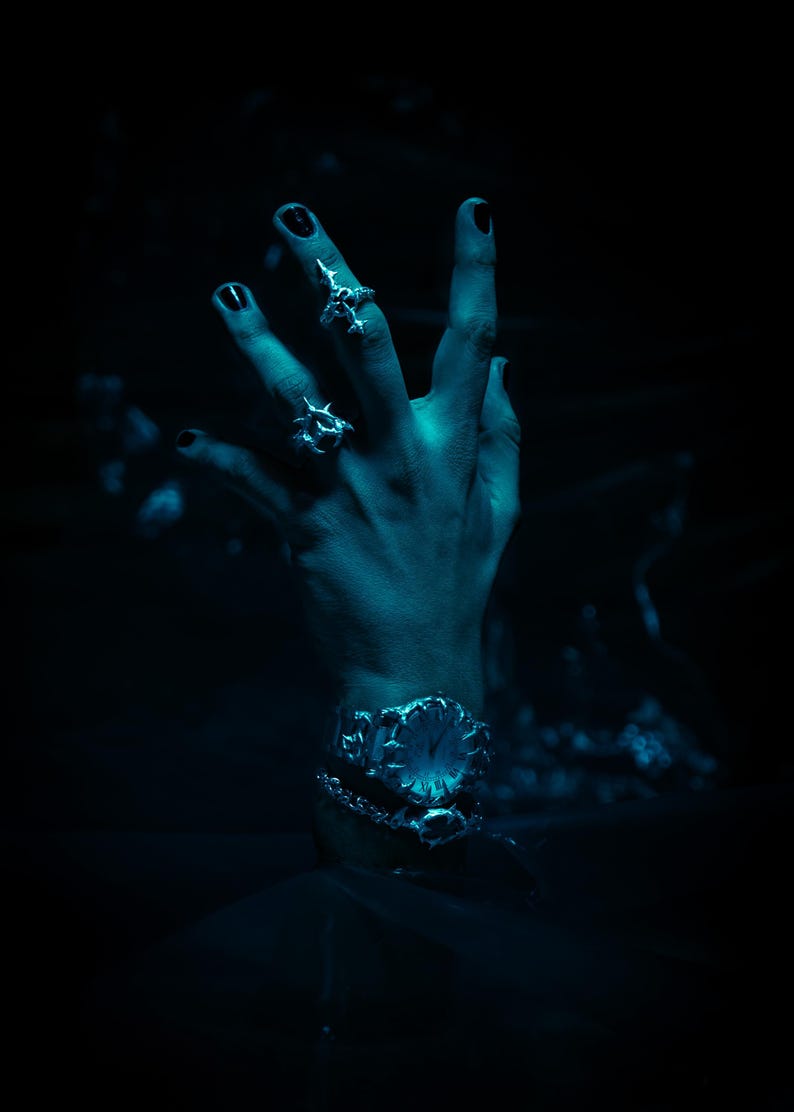 Puede incluir: Una mano con varios anillos de plata y una pulsera de plata con un dise&ntilde;o floral. La mano est&aacute; iluminada con una luz azul sobre un fondo oscuro.