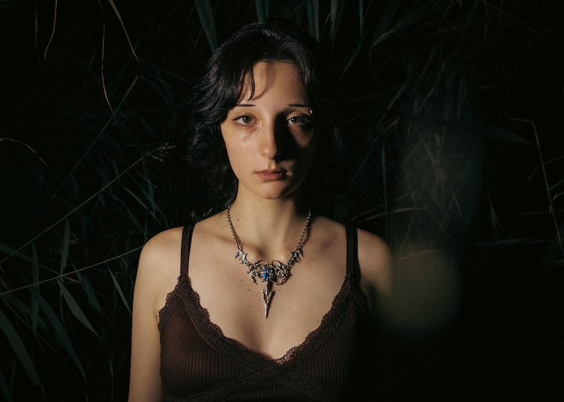 May include: A silver-toned necklace with a central blue stone and ornate, stylized wing-like designs. The necklace is worn over a brown top with lace trim. The background is dark with green foliage.