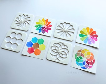 May include: An assortment of white square stencils with various geometric designs, including a spiral, honeycomb, and floral patterns. Adjacent to the stencils are watercolor paintings of the same designs in vibrant rainbow colors. The designs are on a white surface.