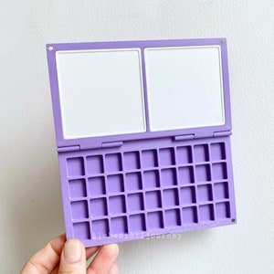 Puede incluir: Una caja de plástico púrpura con dos paneles abatibles blancos y una cuadrícula de 24 cuadrados pequeños.