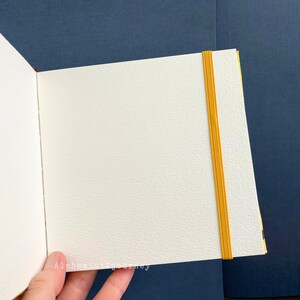 Może przedstawiać: Biały, pusty notes z gumką w kolorze ż&oacute;łtym. Notes jest otwarty na pustej stronie i trzymany w dłoni. Tło to ciemnoniebieska powierzchnia.
