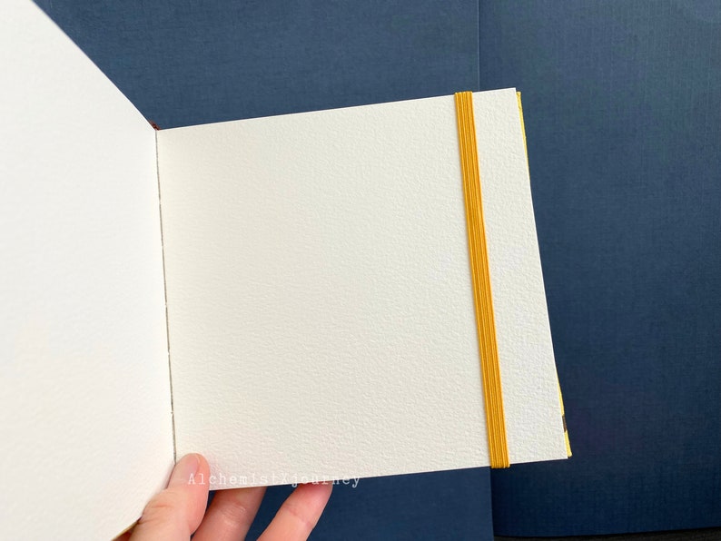 Może przedstawiać: Biały, pusty notes z gumką w kolorze ż&oacute;łtym. Notes jest otwarty na pustej stronie i trzymany w dłoni. Tło to ciemnoniebieska powierzchnia.