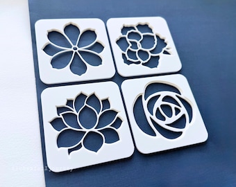 May include: Four white plastic stencils with different flower designs. The stencils are arranged in a square shape on a blue background.