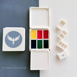 Könnte beinhalten: Ein weißes Aquarellfarbenset mit sechs leuchtenden Farben, darunter Gelb, Rot und Grün, in einem kompakten, klappbaren Etui. Ein separates Etui mit einem blauen Emblem ist ebenfalls vorhanden, zusammen mit mehreren leeren Mischpaletten. Der Text "AlchemistXjourney" ist sichtbar.
