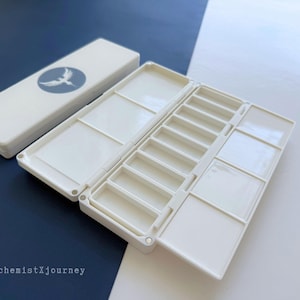 May include: A white plastic watercolor paint box with multiple mixing wells and a closed lid featuring a circular gray logo with a white bird graphic. The text AlchemistXjourney is printed in the lower left corner of the image.