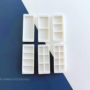 May include: Six white rectangular paint palettes arranged in two rows. The palettes feature varying internal compartment configurations, ranging from a single open well to multiple divided sections. The text AlchemistXjourney is visible.
