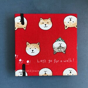 Może przedstawiać: Czerwony notes z tkaniny z czarną gumką. Okładka ma powtarzający się wz&oacute;r kresk&oacute;wkowych ps&oacute;w Shiba Inu i napis "Let's go for a walk!"