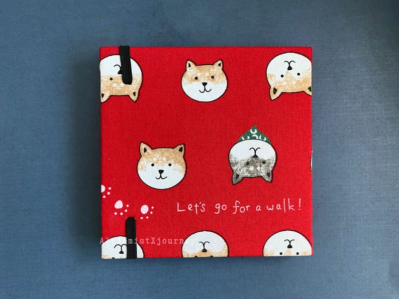 Może przedstawiać: Czerwony notes z tkaniny z czarną gumką. Okładka ma powtarzający się wz&oacute;r kresk&oacute;wkowych ps&oacute;w Shiba Inu i napis "Let's go for a walk!"