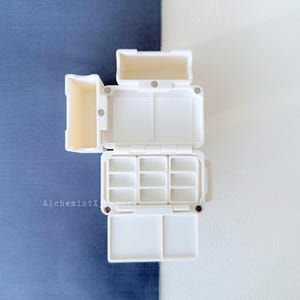 Op de afbeelding: Een witte, open aquarelverfpalet met meerdere compartimenten en een mengruimte. Het palet is rechthoekig en heeft verschillende secties voor het vasthouden van verf. De achtergrond is verdeeld tussen een blauw en wit oppervlak.