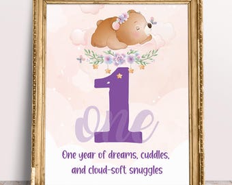 May include: A framed print featuring a sleeping brown bear with a floral crown, a large purple number one, the word one in script, and the text One year of dreams, cuddles, and cloud-soft snuggles against a soft pink background with clouds.