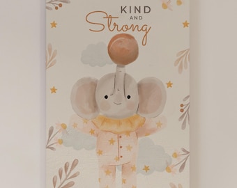 May include: A rectangular wall art print featuring a watercolor illustration of a gray elephant wearing peach-colored pajamas with yellow stars. The text "KIND AND STRONG" is printed above the elephant, surrounded by clouds, stars, and foliage.