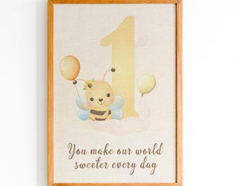 May include: A framed print featuring a large yellow number one, a cartoon bee with blue wings holding an orange balloon, a yellow balloon, and the text "You make our world sweeter every day" in brown script on a light beige textured background.