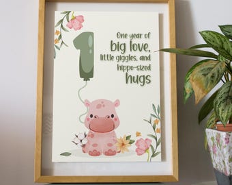 May include: A framed print featuring a light pink cartoon hippo, a green balloon shaped like the number one, and floral accents. The text reads: One year of big love, little giggles, and hippo-sized hugs. The frame is light wood.