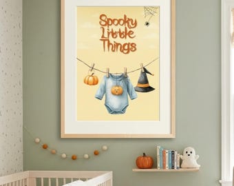 May include: A framed wall art print featuring the text Spooky Little Things in orange letters. A clothesline displays a small orange pumpkin, a light blue baby bodysuit with a pumpkin graphic, and a black witch hat with an orange band.