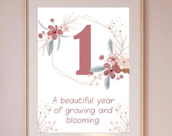 May include: A framed wall art print featuring a large dusty rose number one centered within a thin gold geometric frame. The design is decorated with watercolor flowers and leaves in shades of pink and grey, with the text A beautiful year of growing and blooming printed in a simple font below the number.