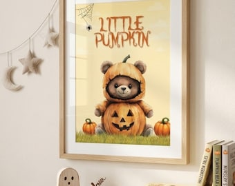 May include: A framed wall art print featuring a brown teddy bear wearing a pumpkin costume with the text Little Pumpkin above it. The illustration includes a spider web, a small spider, and two orange pumpkins on a light yellow background.