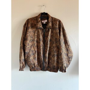 Puede incluir: Una chaqueta de manga larga con cremallera y un estampado animal en tonos marrón y negro que combina manchas de leopardo y rayas de cebra. La prenda cuenta con cuello alto, cremallera frontal completa y puños y dobladillo elásticos.