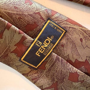May include: A Fendi silk tie with a repeating leaf pattern in shades of brown and red. The tie features a navy blue label with the Fendi logo and the words "Made in Italy".