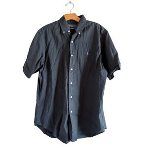 Puede incluir: Camisa abotonada de manga corta azul marino oscuro con cuello abotonado. La camisa tiene un pequeño logotipo morado en el pecho izquierdo. Colgada de una percha de madera.