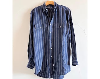 May include: Navy blue long-sleeve shirt with vertical white stripes. The shirt has two chest pockets and a button-down front. The shirt is hanging on a wooden hanger.