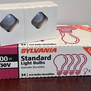 May include: Two boxes of Sylvania Standard Light Bulbs. One box is white with two bulbs visible through diamond-shaped cutouts. The other box is a case pack with the text "100W 130V" and "24 A19 Bulbs".