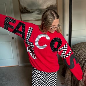 May include: A red sweatshirt with the word "FALCONS" in patchwork letters. The letters are black and white polka dot, checkered, and striped. The sweatshirt has a team logo on the front and is paired with black and white checkered pants.